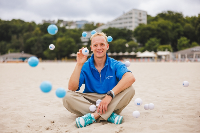 Student Council | WSB Merito Gdańsk University: Grafika główna: samorząd studencki Gdańsk, student siedzi na plaży i trzyma piłeczkę z logo WSB Merito