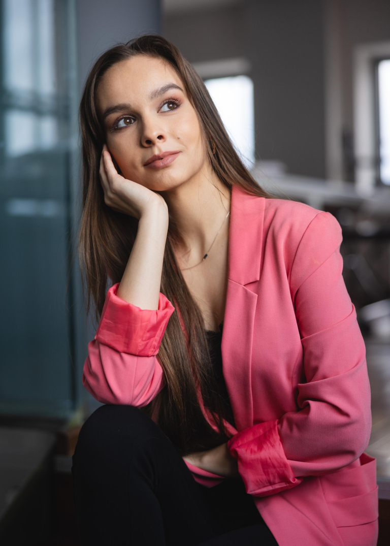 studentka z długimi włosami i w marynarce w kolorze łososiowym, siedzi na sofie, patrzy za okno, głowę podpiera na ręce