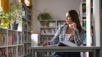Wyniki wyszukiwania | Uniwersytet WSB Merito w Gdańsku: Studentka w bibliotece siedzi przy stoliku przy książce i patrzy przez okno