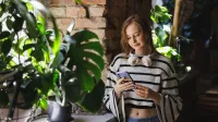 Digital Marketing | Studia II stopnia ze studiami podyplomowymi  | Uniwersytet WSB Merito w Gdańsku: Studentka z komórką stoi przy oknie przy zielonym parapecie