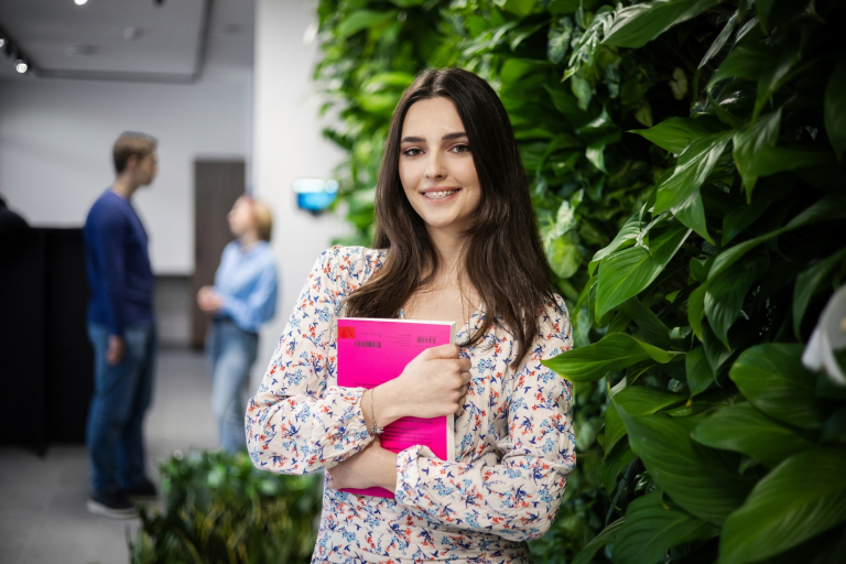 Psychologia w marketingu | Studia II stopnia ze studiami podyplomowymi  | Uniwersytet WSB Merito w Gdańsku: Studentka z książką na tle zielonej ściany z liści