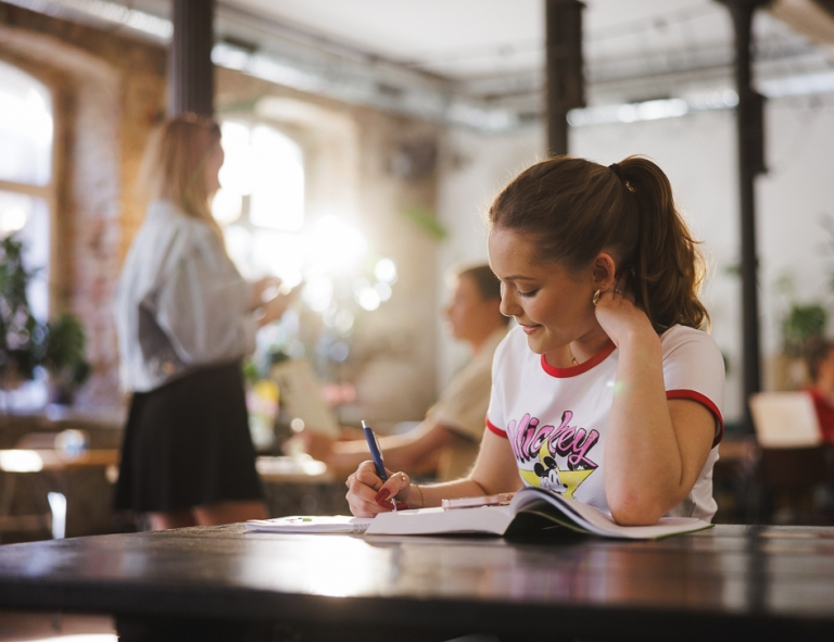 Copywriting | Studia II stopnia w Gdańsku | Uniwersytet WSB Merito w Gdańsku: Studentka notuje coś w zeszycie w przestrzeni kawiarni 