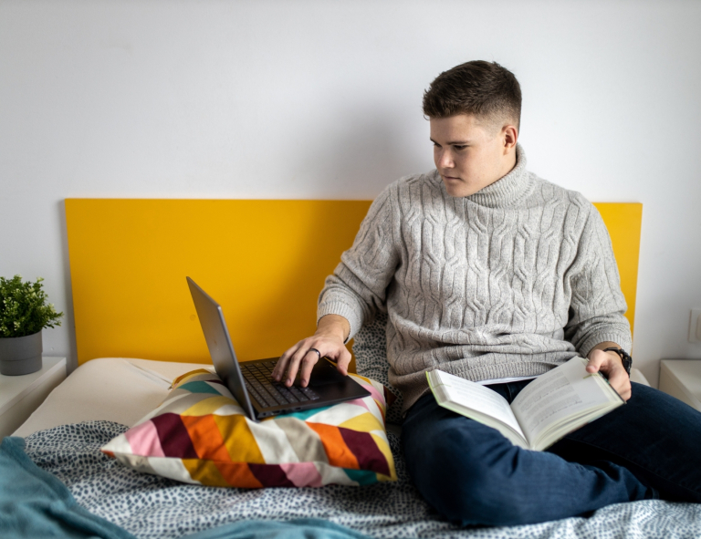 Informatyka | Studia I stopnia w Gdańsku | Uniwersytet WSB Merito w Gdańsku: Student siedzi w łóżku z laptopem i ma na kolanach książkę