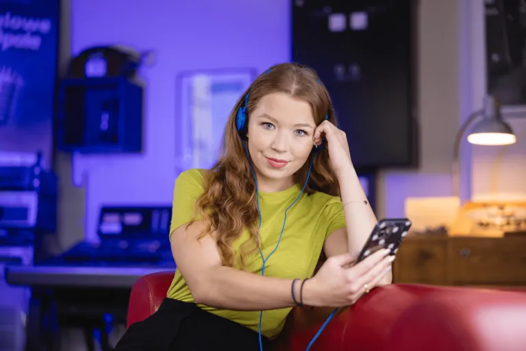 Produkcja audiowizualna w mediach cyfrowych | Studia I stopnia w Gdańsku | Uniwersytet WSB Merito w Gdańsku: Na zdjęciu znajduje się młoda dziewczyna, która ma w ręku komórkę oraz nosi niebieskie słuchawki. Ma ona rudawe włosy i lekko się uśmiecha