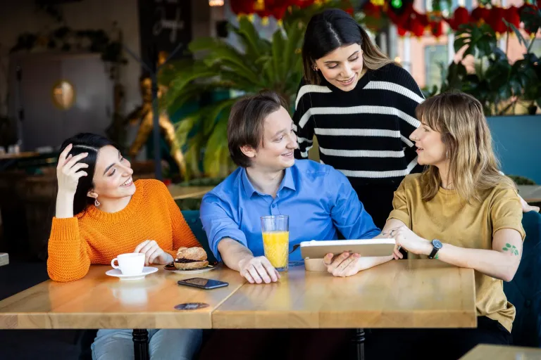 Edukacja Dorosłych z Animacją Społeczno-Kulturalną | Studia II stopnia ze studiami podyplomowymi  | Uniwersytet WSB Merito w Gdańsku: Studenci siedzą przy biurku w kawiarni i rozmawiają, jedna z osób trzyma tablet
