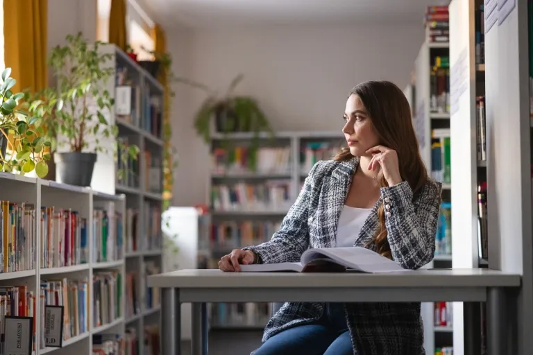 Zarządzanie przedsiębiorstwem | Studia II stopnia w Gdańsku | Uniwersytet WSB Merito w Gdańsku: Studentka w bibliotece siedzi przy stoliku przy książce i patrzy przez okno