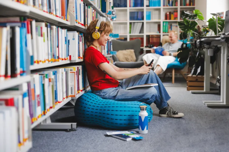 Digital Marketing | Studia II stopnia w Gdańsku | Uniwersytet WSB Merito w Gdańsku: Studentka z książką na kolanach, słuchawkami na uszach i z komórką w ręku we wnętrzu biblioteki