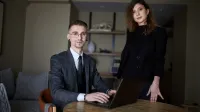Tester oprogramowania dla aplikacji mobilnych i serwerowych z zastosowaniem AI | Studia podyplomowe w Gdyni | Uniwersytet WSB Merito Gdynia: Zdjęcie osób w średnim wieku. Mężczyzna w garniturze siedzi przy laptopie, obok niego stoi kobieta ubrana na czarno