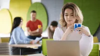 Rozwój produktów w małej firmie | Studia podyplomowe w Gdyni | Uniwersytet WSB Merito Gdynia: Zdjęcie kobiety siedzącej w przestrzeni kawiarni przy laptopie. Z tyłu widać innych ludzi. Ma ona na sobie jasne ubrania i trzyma kubek z logo Merito