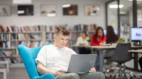Zarządzanie nieruchomościami i homestaging | Studia II stopnia w Gdyni | Uniwersytet WSB Merito Gdynia: Student w przestrzeni biblioteki siedzi na fotelu i na kolanach trzyma laptopa