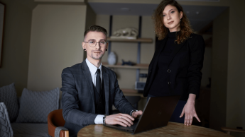 Tester oprogramowania dla aplikacji mobilnych i serwerowych z zastosowaniem AI | Studia podyplomowe w Gdyni | Uniwersytet WSB Merito Gdynia: Zdjęcie osób w średnim wieku. Mężczyzna w garniturze siedzi przy laptopie, obok niego stoi kobieta ubrana na czarno