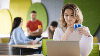 Rozwój produktów w małej firmie | Studia podyplomowe w Gdyni | Uniwersytet WSB Merito Gdynia: Zdjęcie kobiety siedzącej w przestrzeni kawiarni przy laptopie. Z tyłu widać innych ludzi. Ma ona na sobie jasne ubrania i trzyma kubek z logo Merito