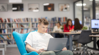 Zarządzanie nieruchomościami i homestaging | Studia II stopnia w Gdyni | Uniwersytet WSB Merito Gdynia: Student w przestrzeni biblioteki siedzi na fotelu i na kolanach trzyma laptopa