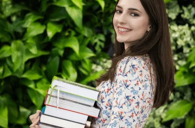 Biblioteka | Uniwersytet WSB Merito Gdynia: Zdjęcie młodej studentki z aparatem na zębach. Ma brązowe włosy do ramion i delikatną białą sukienkę w małe kwiatki. Trzyma w ręku stertę książek. Pozuje na tle zielonej ściany z kwiatami