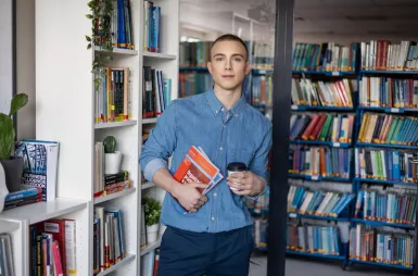 Biblioteka | Uniwersytet WSB Merito Gdynia: Student z książkami i kawą w ręku w przestrzeni biblioteki
