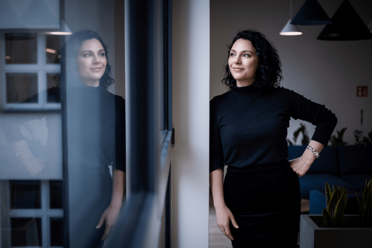 Program firma | Uniwersytet WSB Merito Gdynia: Portret kobiety w czarnych kręconych włosach ubranej również w ciemne barwy. Opiera się ona o ścianę, a drugą rękę opiera o biodro, uśmiecha się. W oknie widać jej odbicie