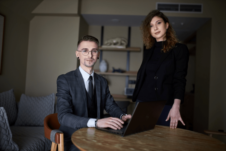 Zarządzanie środowiskiem i ESG z certyfikatem Audytora Wewnętrznego ISO 14001 | Studia podyplomowe w Gdyni | Uniwersytet WSB Merito Gdynia: Zdjęcie osób w średnim wieku. Mężczyzna w garniturze siedzi przy laptopie, obok niego stoi kobieta ubrana na czarno
