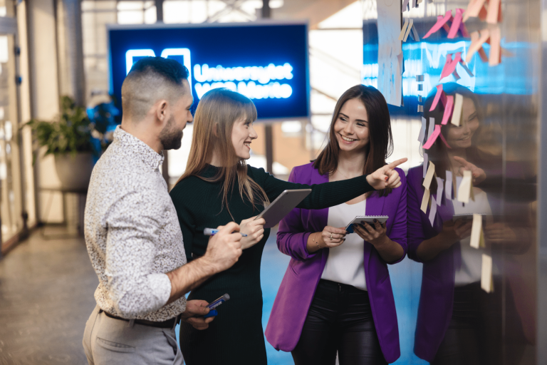 Program firma | Uniwersytet WSB Merito Gdynia: Zdjęcie przedstawiające dwie studentki i studenta pracującego w grupie. Stoją oni przy karteczkach i coś omawiają. Ubrani są biznesowo. Z tyłu widać neon Uniwersytetu WSB Merito