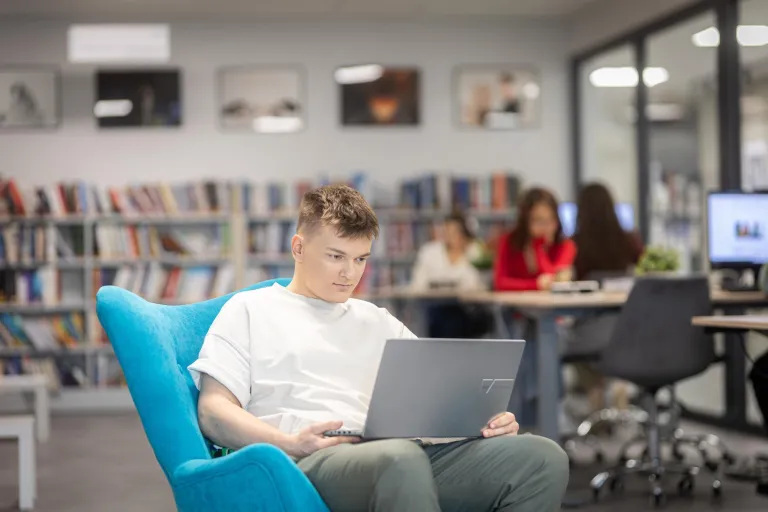 Informatyka | Studia I stopnia w Gdyni | Uniwersytet WSB Merito Gdynia: Student w przestrzeni biblioteki siedzi na fotelu i na kolanach trzyma laptopa