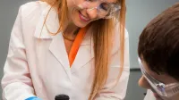 Zarządzanie i kontrola w podmiotach leczniczych | Uniwersytet WSB Merito Łódź: Kobieta w laboratorium w białym fartuchu i okularach ochronnych pokazuje dziecku, jak korzystać z miernika pH, wykonując doświadczenie przy stole z próbkami.