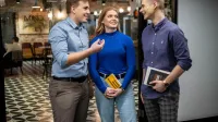 Zarządzanie zespołami rozproszonymi | Studia podyplomowe w Łodzi | Uniwersytet WSB Merito Łódź: Trójka studentów rozmawia ze sobą w przestrzeni coworkingowej, trzymając książki i uśmiechając się podczas dyskusji.