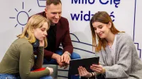 Rachunkowość przedsiębiorstw i kontrola finansowa | Studia II stopnia w Łodzi | Uniwersytet WSB Merito Łódź: Troje młodych ludzi wspólnie ogląda coś na tablecie, siedząc w nowoczesnym wnętrzu z napisem „Miło Cię widzieć” w tle.