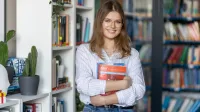 Trener języków obcych | Uniwersytet WSB Merito Łódź: Młoda kobieta w okularach, trzymająca podręcznik w bibliotece, uśmiecha się stojąc przy regale z książkami.