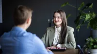 Energetyka wiatrowa (Onshore & Offshore) | Uniwersytet WSB Merito Łódź: Kobieta w okularach i eleganckim stroju uśmiecha się podczas rozmowy z mężczyzną w jasnoniebieskiej koszuli przy biurku w nowoczesnym biurze.