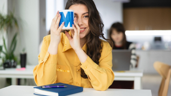 Business English | Studia podyplomowe w Łodzi | Uniwersytet WSB Merito Łódź: Uśmiechnięta dziewczyna trzyma kubek "Merito" przy twarzy.