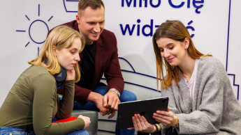 Rachunkowość przedsiębiorstw i kontrola finansowa | Studia II stopnia w Łodzi | Uniwersytet WSB Merito Łódź: Troje młodych ludzi wspólnie ogląda coś na tablecie, siedząc w nowoczesnym wnętrzu z napisem „Miło Cię widzieć” w tle.