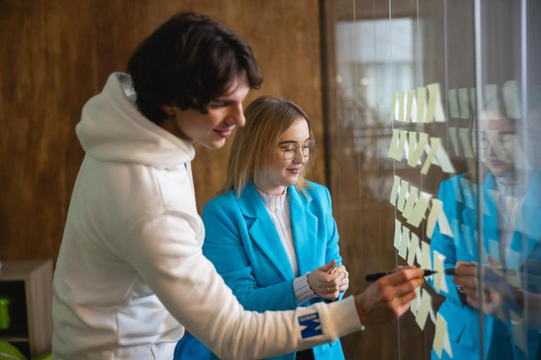 Management | Studia I stopnia w Łodzi | Uniwersytet WSB Merito Łódź: Dwoje studentów pracuje wspólnie przy szklanej tablicy z karteczkami, planując i omawiając elementy projektu.