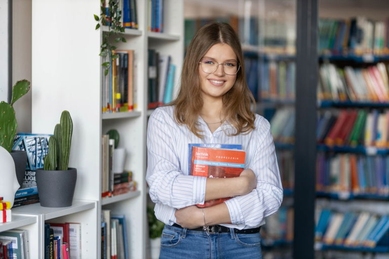 Trening menedżerski, studia podyplomowe: Młoda kobieta w okularach, trzymająca podręcznik w bibliotece, uśmiecha się stojąc przy regale z książkami.