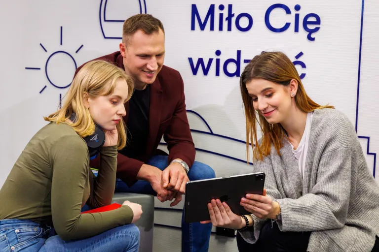 AI w zarządzaniu | Studia II stopnia ze studiami podyplomowymi  | Uniwersytet WSB Merito Łódź: Troje młodych ludzi wspólnie ogląda coś na tablecie, siedząc w nowoczesnym wnętrzu z napisem „Miło Cię widzieć” w tle.