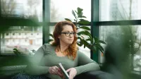 Główna grafika: Opole - studia podyplomowe - Terapia uzależnień - uczestniczka studiów podyplomowych siedzi na kanapie z książką w rękach i spogląda za okno
