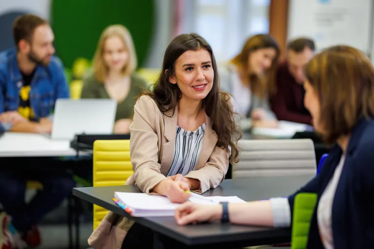 Komunikacja wizerunkowa | Studia I stopnia w Opolu | Uniwersytet WSB Merito Opole: Dwie studentki rozmawiają o projekcie, w tle inni studenci
