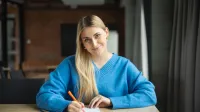 Psychologia negocjacji i mediacji | Studia I stopnia w Poznaniu | Uniwersytet WSB Merito w Poznaniu: kobieta w niebieskim swetrze siedzi przy stole i z uśmiechem sporządza notatki