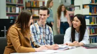 Zarządzanie gospodarką komunalną | Studia II stopnia ze studiami podyplomowymi  | Uniwersytet WSB Merito w Poznaniu: Troje studentów uczy się wspólnie przy stole w bibliotece, śmiejąc się