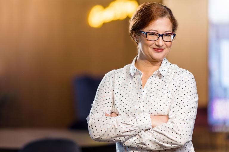 Oferty pracy | Uniwersytet WSB Merito w Poznaniu: Kobieta w okularach uśmiecha się, stojąc z założonymi rękami w nowoczesnym wnętrzu.