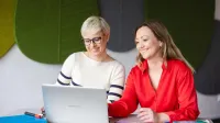 SAP S/4HANA – nowoczesny system ERP | Studia podyplomowe w Szczecinie | Uniwersytet WSB Merito Szczecin: Dwie kobiety pracują przy laptopie, uśmiechając się i omawiając materiały.