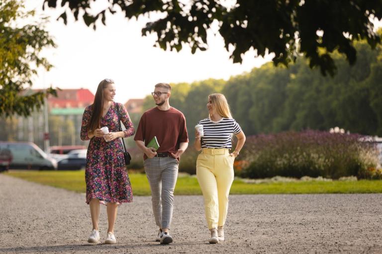 Studia i praktyki za granicą | Uniwersytet WSB Merito Szczecin: Troje studentów Uniwersytetu WSB Merito Szczecin spaceruje po parku, trzymając kawę i rozmawiając.