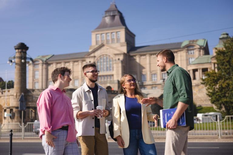 Turystyka i rekreacja | Studia I stopnia w Szczecinie | Uniwersytet WSB Merito Szczecin: Czworo studentów rozmawia na tle gmachu Wałów Chrobrego w słoneczny dzień.