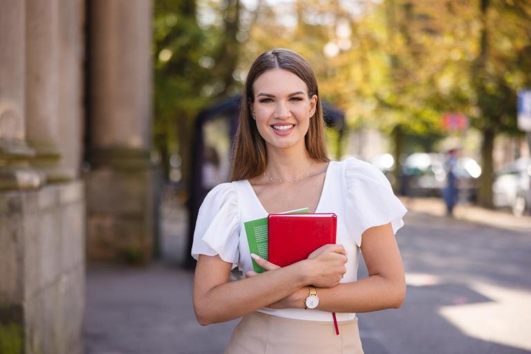 Pedagogika przedszkolna i wczesnoszkolna | Studia jednolite magisterskie w Szczecinie | Uniwersytet WSB Merito Szczecin: Studentka stoi na tle miejskiej ulicy z zeszytami w rękach, uśmiechając się do obiektywu.