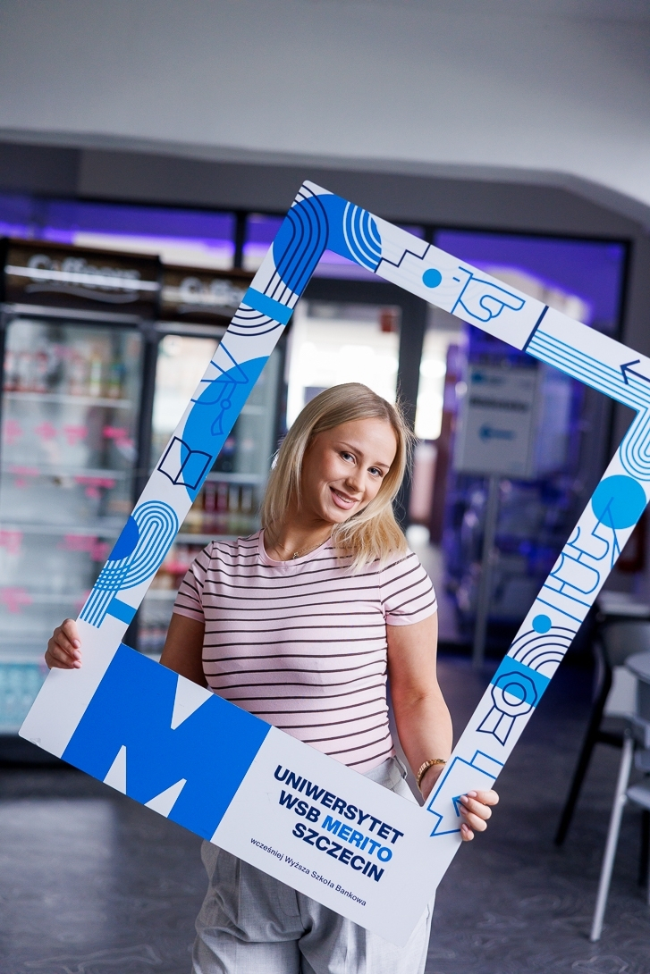 "Potrafię się obronić" – sprawdź, jak ogarnąć stres przed obroną! 🎓 | Aktualności | Uniwersytet WSB Merito Szczecin: Dziewczyna pozuje z dużą ramką uczelni.