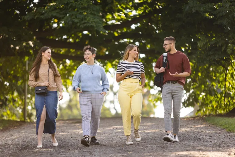 Zarządzanie projektami | Studia I stopnia w Szczecinie | Uniwersytet WSB Merito Szczecin: Czworo studentów spaceruje alejką w parku, rozmawiając i uśmiechając się.
