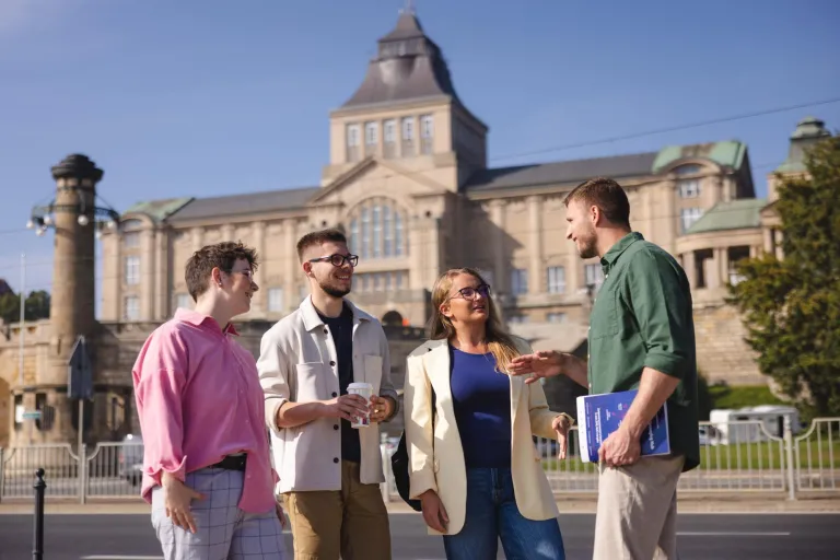 Psychologia w zarządzaniu | Studia II stopnia ze studiami podyplomowymi  | Uniwersytet WSB Merito Szczecin: Czworo studentów rozmawia na tle gmachu Wałów Chrobrego w słoneczny dzień.