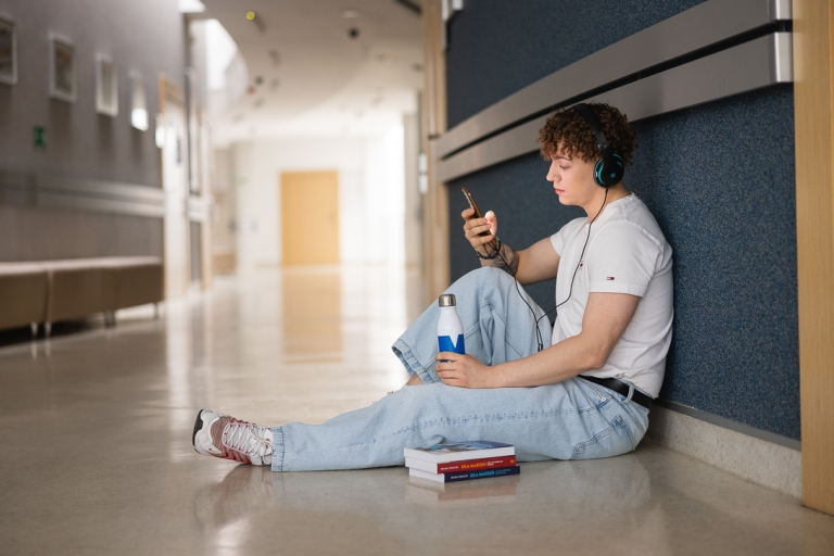 Certyfikaty językowe | Uniwersytet WSB Merito w Toruniu: Student WSB Merito siedzi na korytarzu uczelni oparty o ścianę, przegląda telefon, obok niego na podłodze leży kilka książek