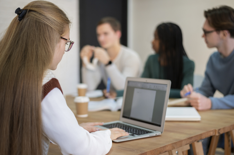 Cloud DevOps Engineer Expert | Studia podyplomowe we Wrocławiu | Uniwersytet WSB Merito we Wrocławiu: Kobieta pracuje przy komputerze, w tle grupa osób z książkami